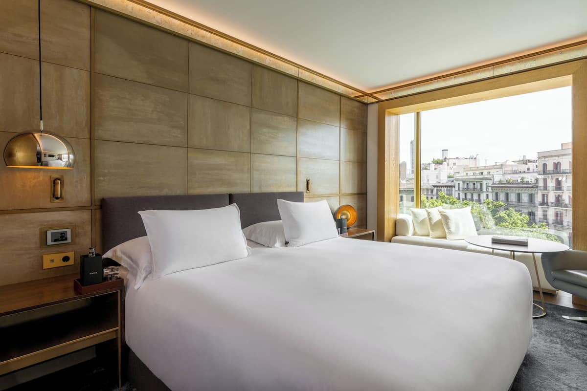 A modern hotel room with a large bed, a bedside table, a lamp, and a view of the city through large windows.
