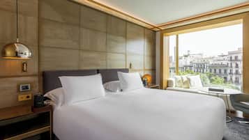 A modern hotel room with a large bed, a bedside table, a lamp, and a view of the city through large windows.