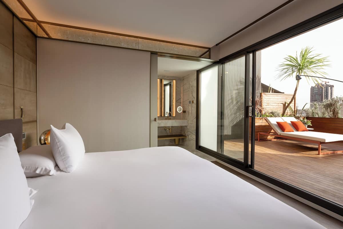 A modern hotel room with a large bed, a bathroom with a mirror, and a balcony with a sun lounger.
