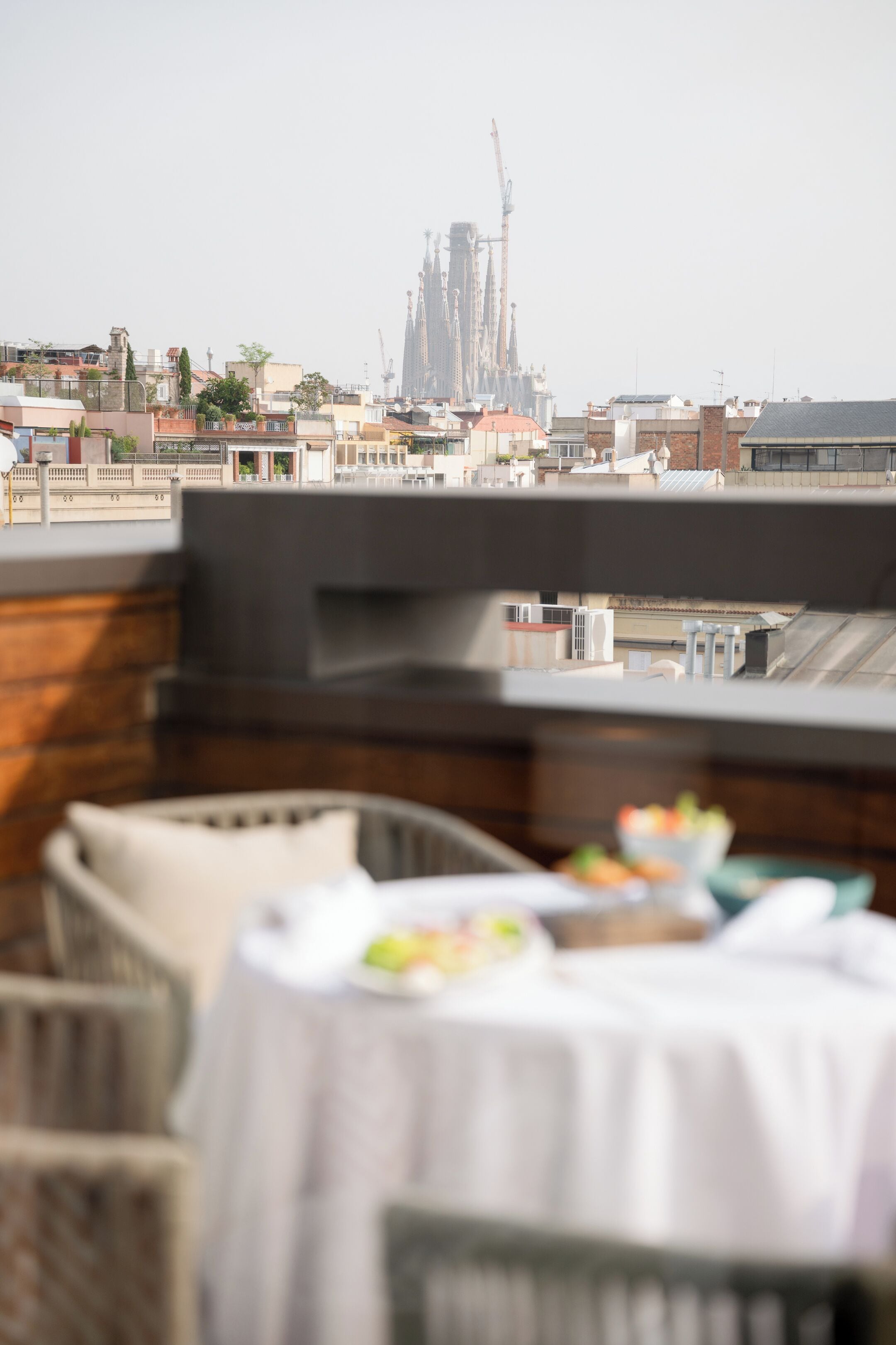 A rooftop terrace with a view of a large building in the distance, a table with a white tablecloth, and a wicker chair.