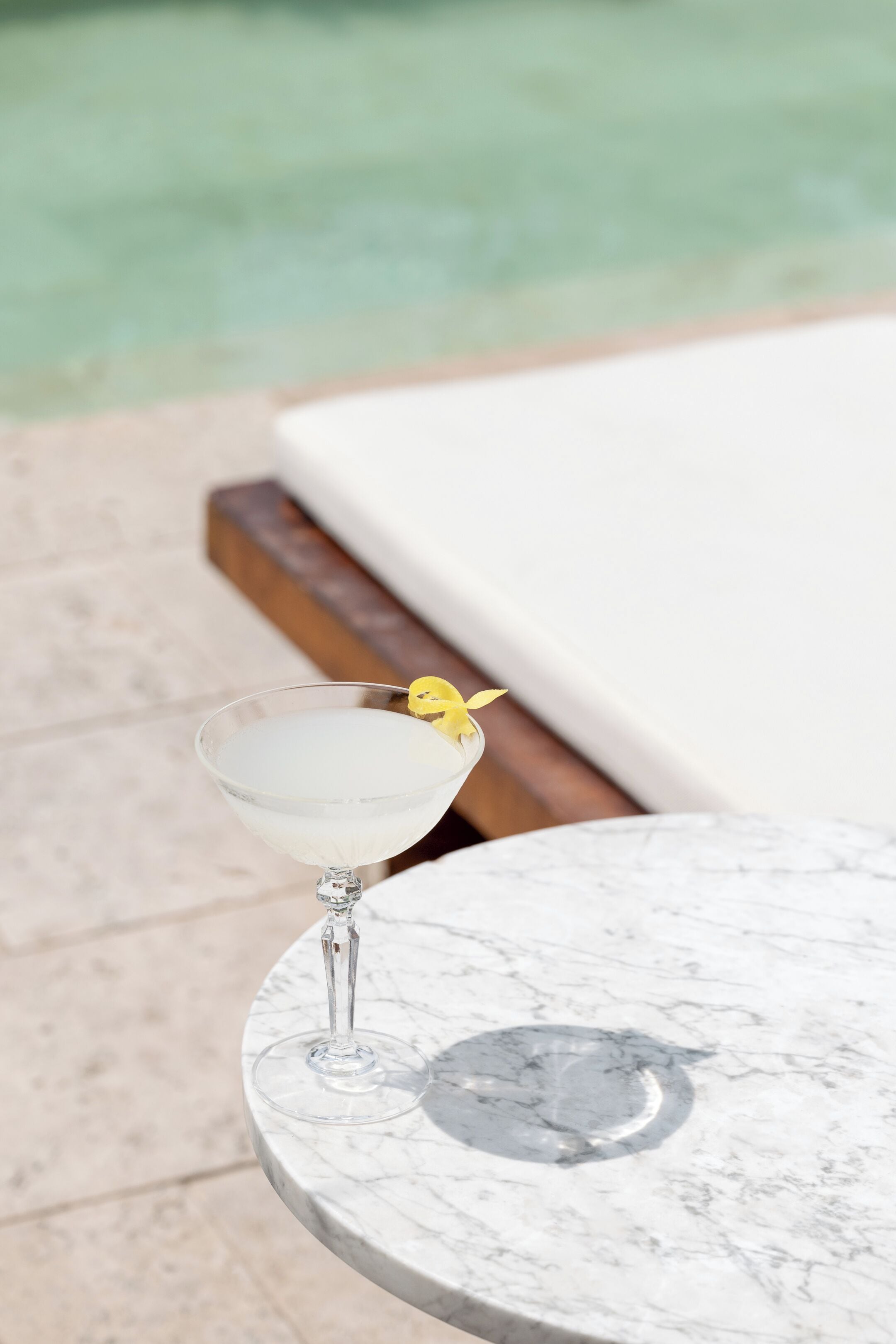 A martini glass with a yellow garnish on a marble table by a poolside lounge area.