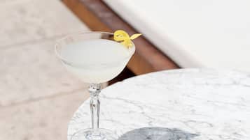 A martini glass with a yellow garnish on a marble table by a poolside lounge area.