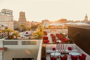 A rooftop terrace with outdoor seating, palm trees, and a cityscape view.