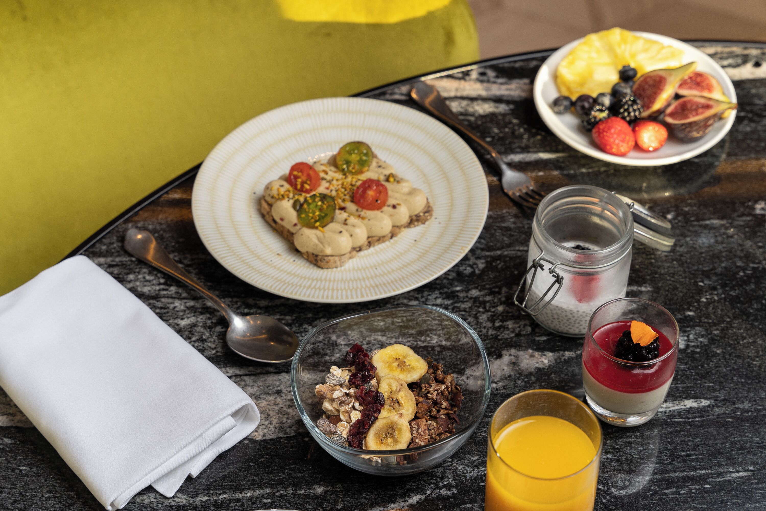 Un desayuno con una fuente de fruta, un tazón de cereal, un vaso de jugo de naranja y un postre pequeño sobre una mesa.