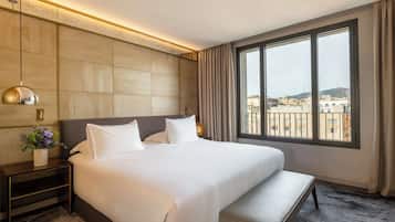 Habitación de hotel con una cama grande, una mesita de noche con lámpara, un banco y un ventanal con vistas a la ciudad.