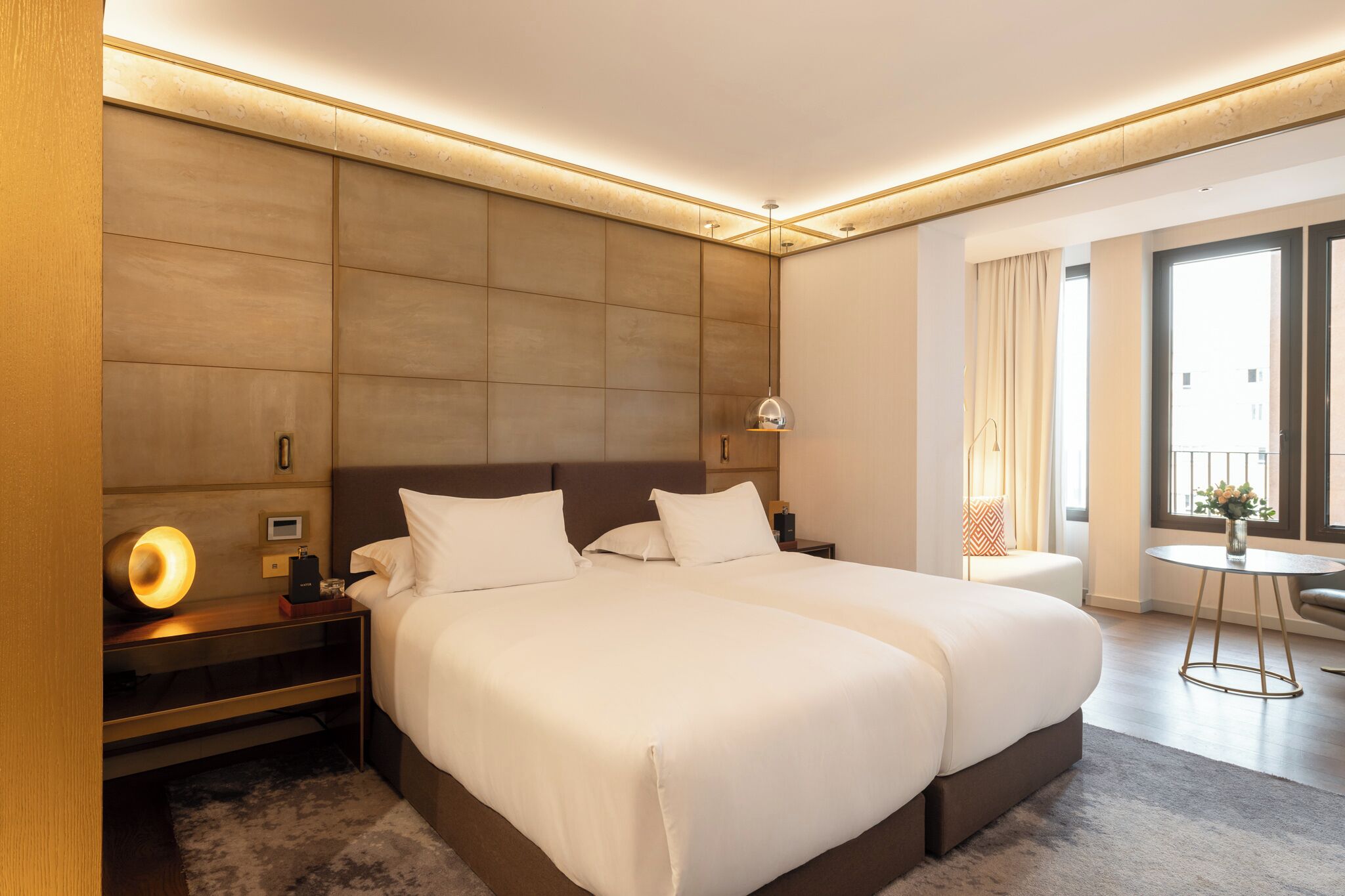 Habitación de hotel con dos camas, una mesita de noche con lámpara, una pequeña mesa con un florero y vistas al exterior a través de una ventana.
