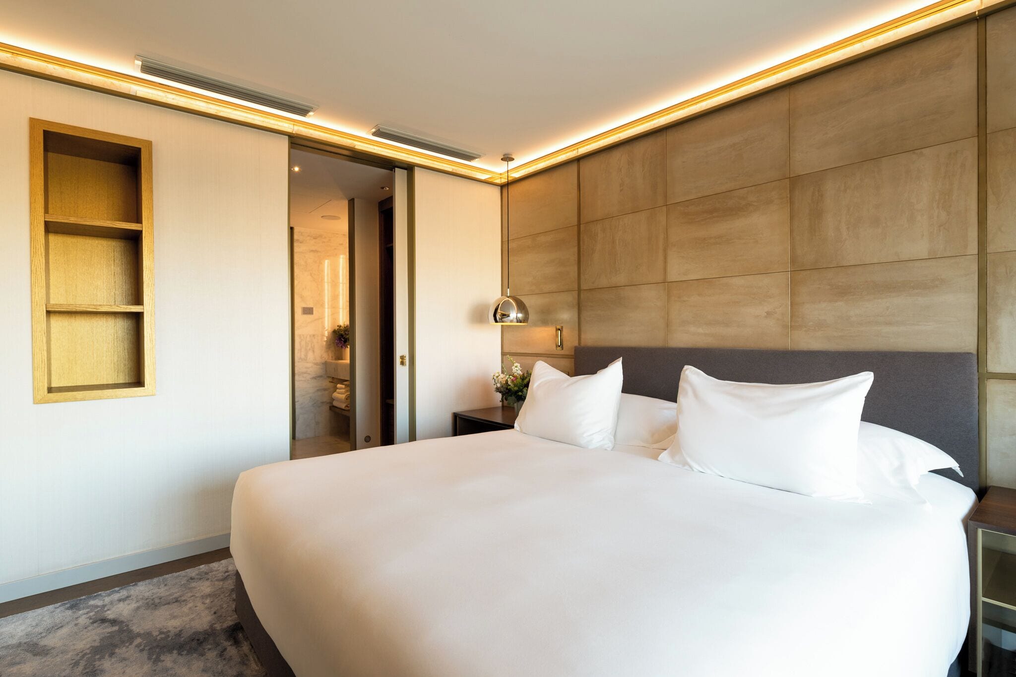 Una habitación de hotel moderna con una cama grande, una mesita de noche con lámpara y estanterías integradas.