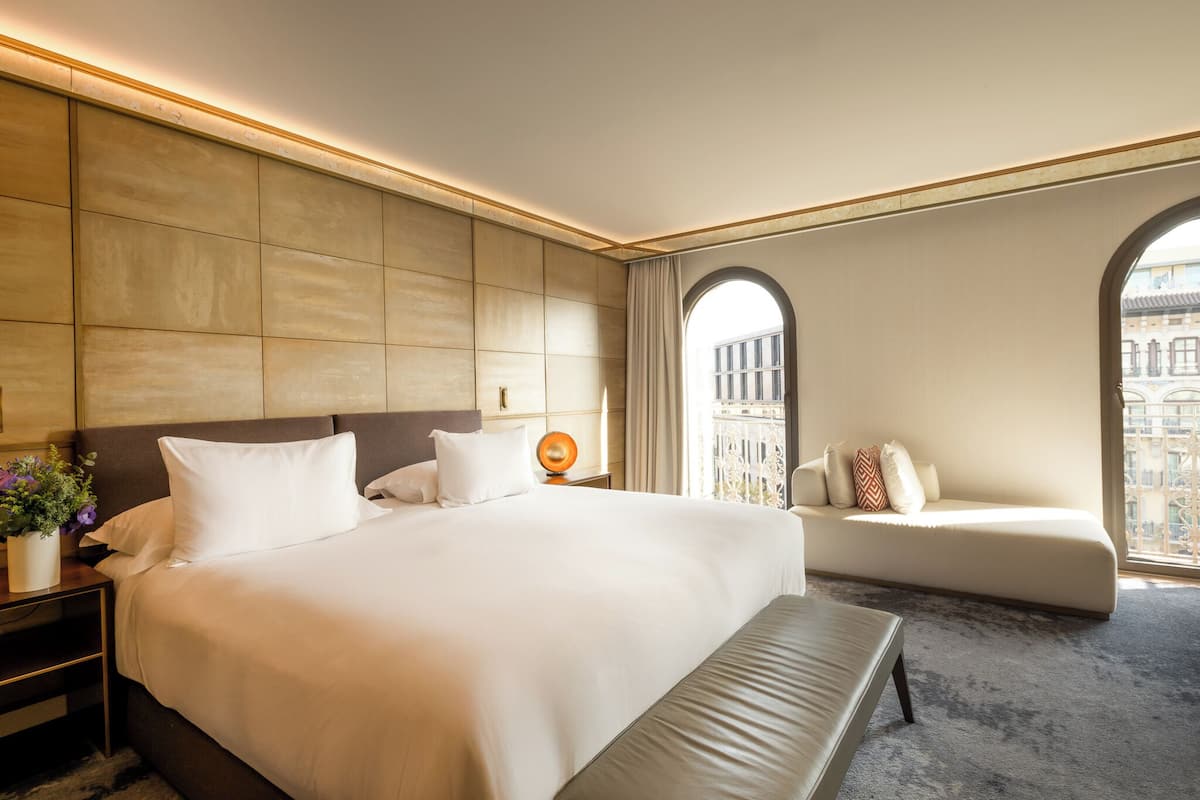 A hotel room with a large bed, a sofa, a nightstand with a lamp, and a view of buildings through arched windows.