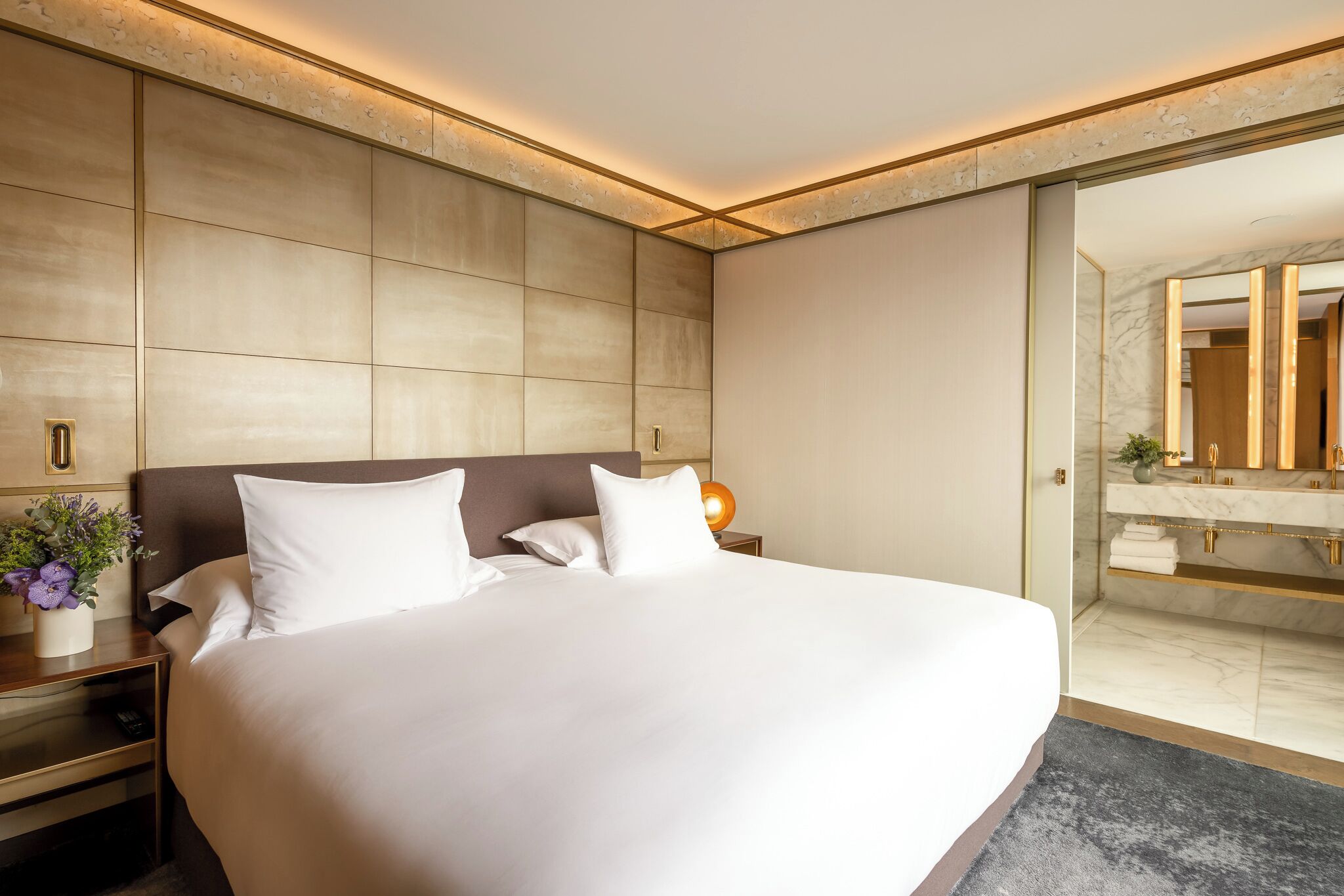 A modern hotel room with a large bed, a nightstand with a lamp, a small table with a vase of flowers, and a bathroom with a marble vanity and large mirror.