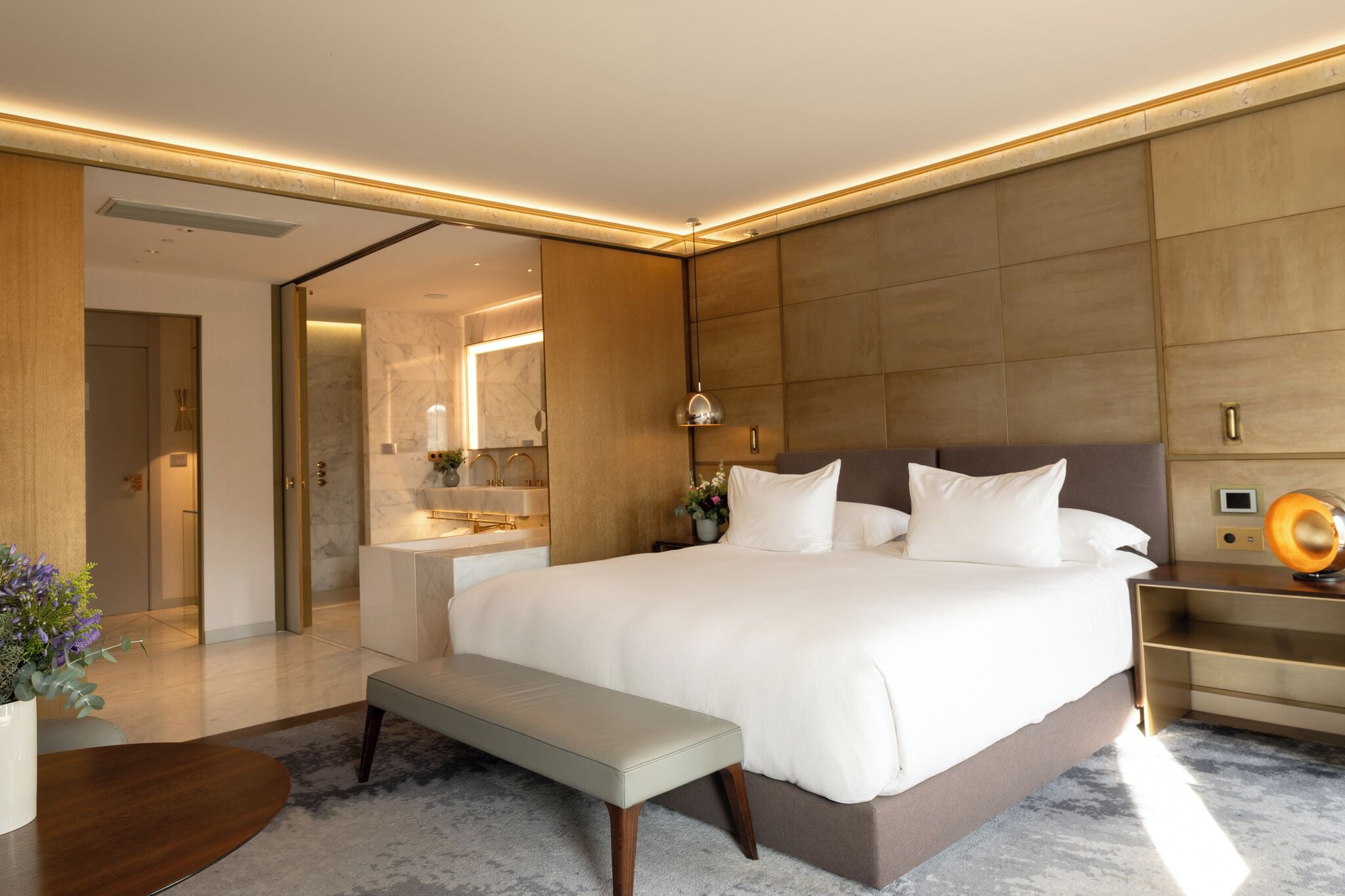 A modern hotel room with a large bed, a bench, a bedside table with a lamp, and a bathroom visible through an open door.