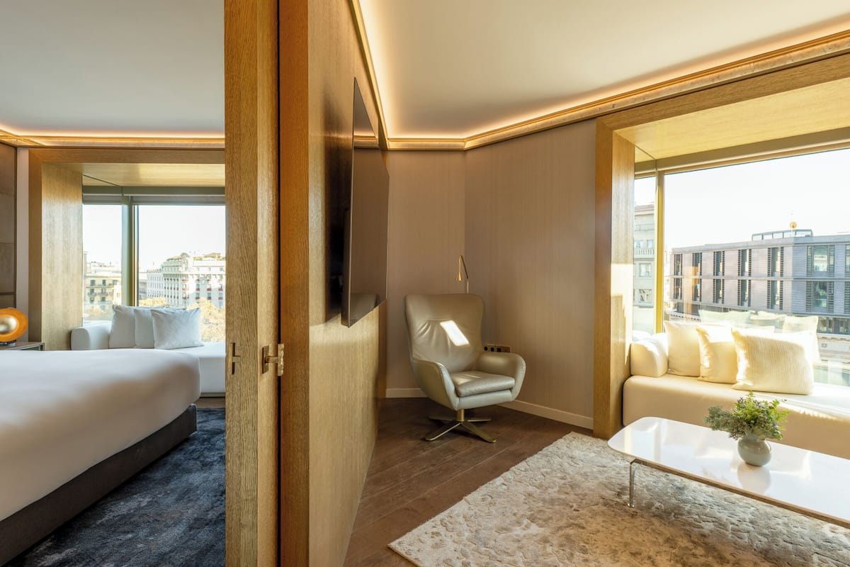 A modern hotel room with a large bed, a seating area with a sofa and armchairs, a flat-screen TV mounted on the wall, and a large window offering a city view.