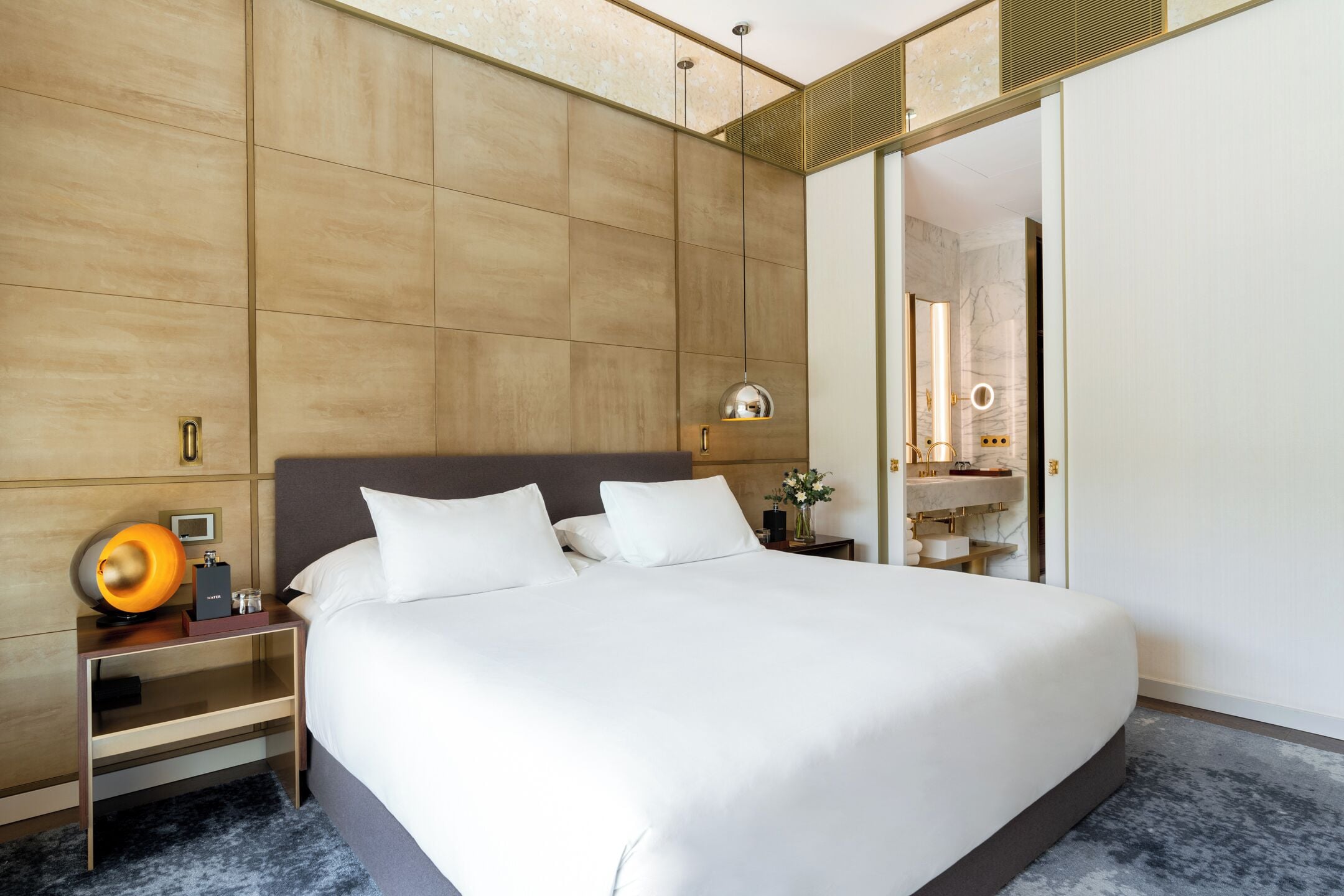 Habitación de hotel moderna con una cama grande, una mesita de noche con lámpara y un baño a la vista a través de una puerta abierta.