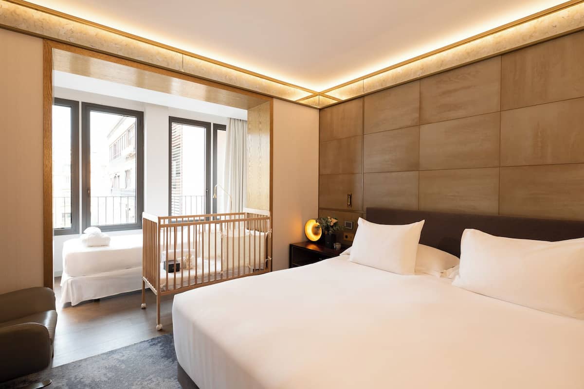 A hotel room with a large bed, a separate seating area with a chair, a small table with a lamp, and a view of the city through large windows.