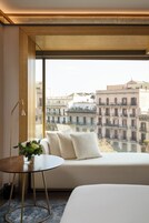 A modern hotel room with a large window, a round wooden table, and a sofa with cushions.