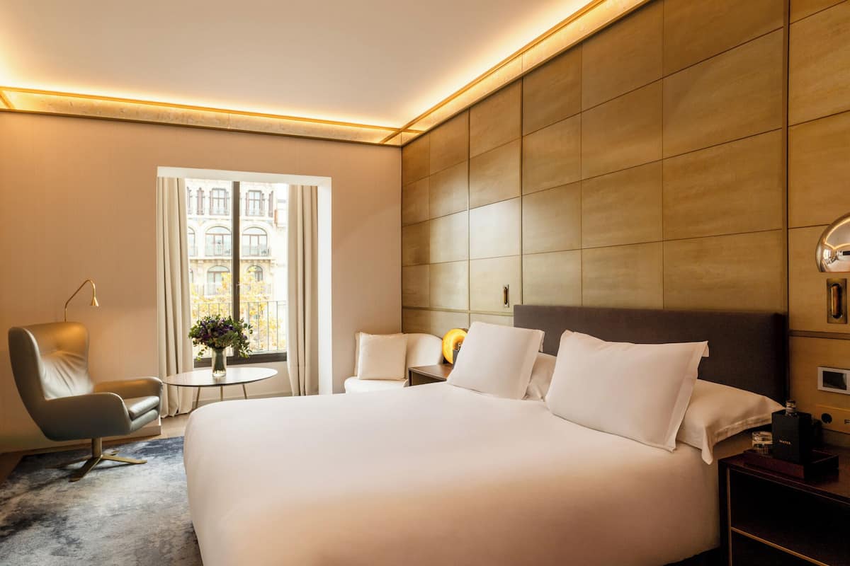 A hotel room with a large bed, a sitting area with a chair and a small table, a nightstand with a lamp, and a view of a building through a window.