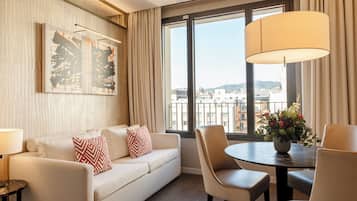 A hotel room with a sofa, a dining table with chairs, a lamp, a window with a view of buildings, and a painting on the wall.