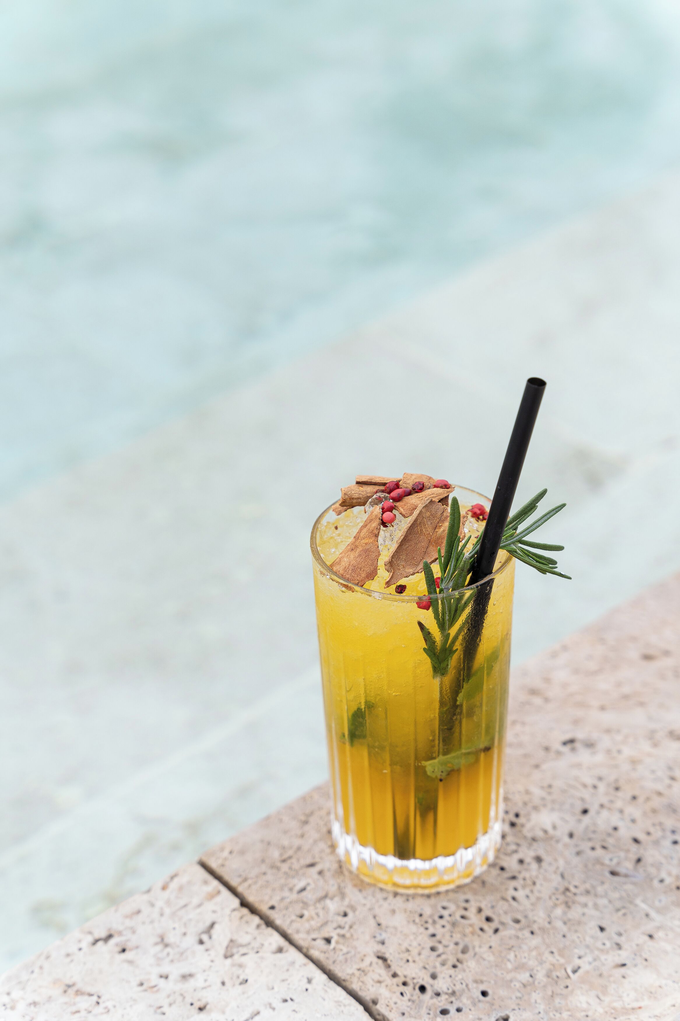 A tall glass with a straw, containing a yellow beverage, garnished with herbs and spices, placed on a textured surface with a blurred background.