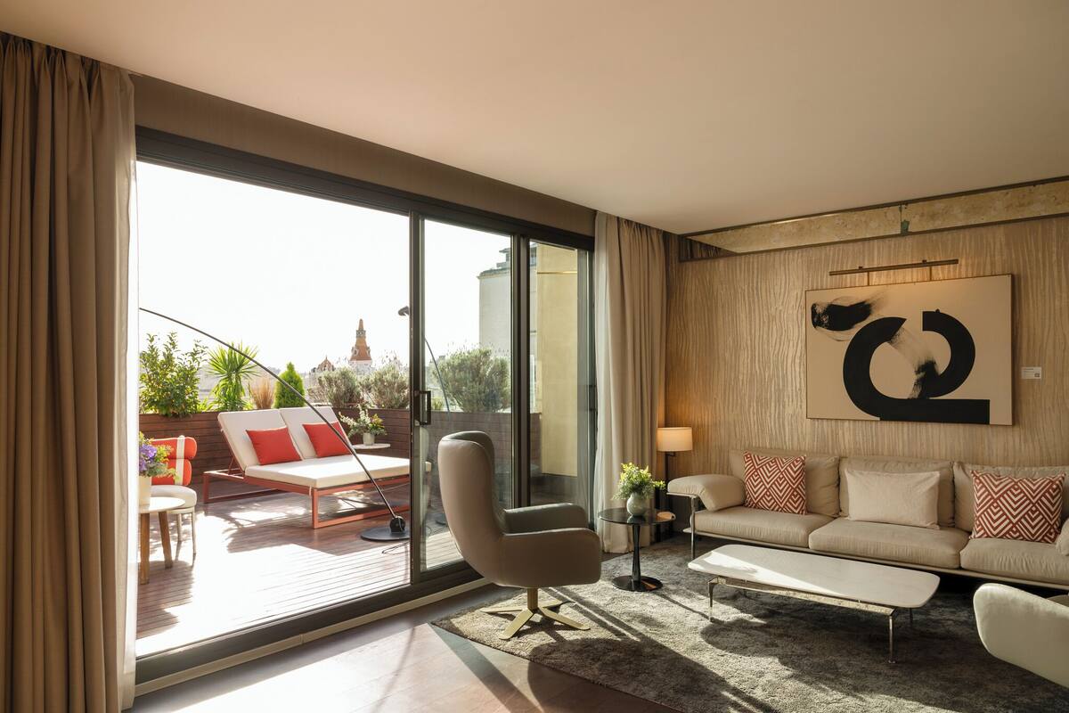 A modern living room with a sofa, armchair, and coffee table. There is a large window with curtains, a painting on the wall, and a view of the city.