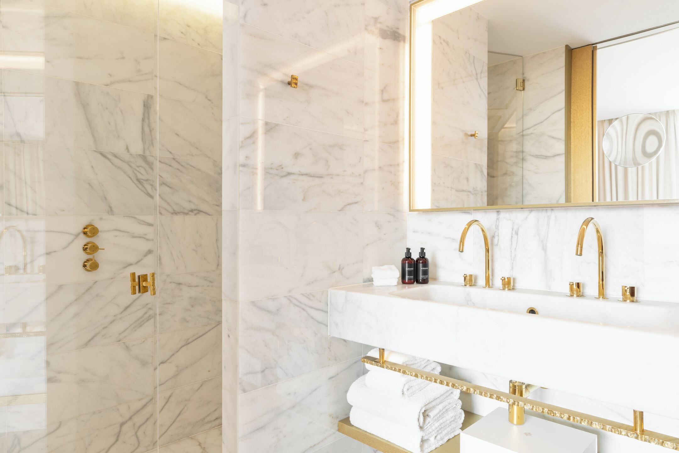 Een moderne badkamer met een marmeren wand, een grote spiegel, gouden kraanwerk en een witte wastafel.