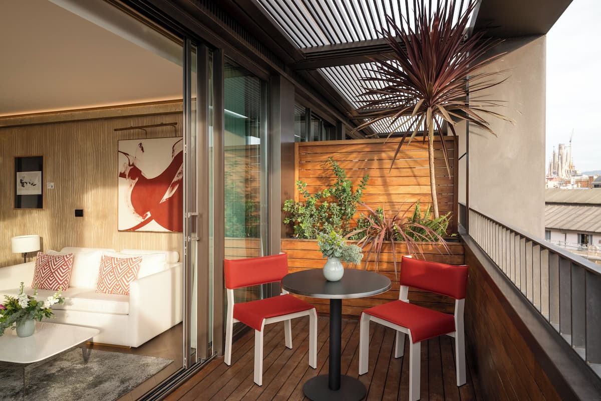 A modern balcony with a wooden floor, a white sofa with patterned cushions, a round table with red chairs, and a potted plant.
