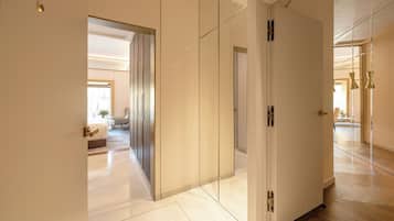 A modern hotel corridor with a wooden floor, a white door, and a view into a well-lit room with a bed and a small table.