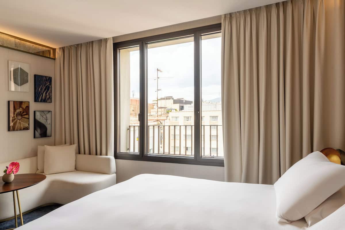 A hotel room with a large bed, a sofa, a small table with a vase, and a window with curtains.