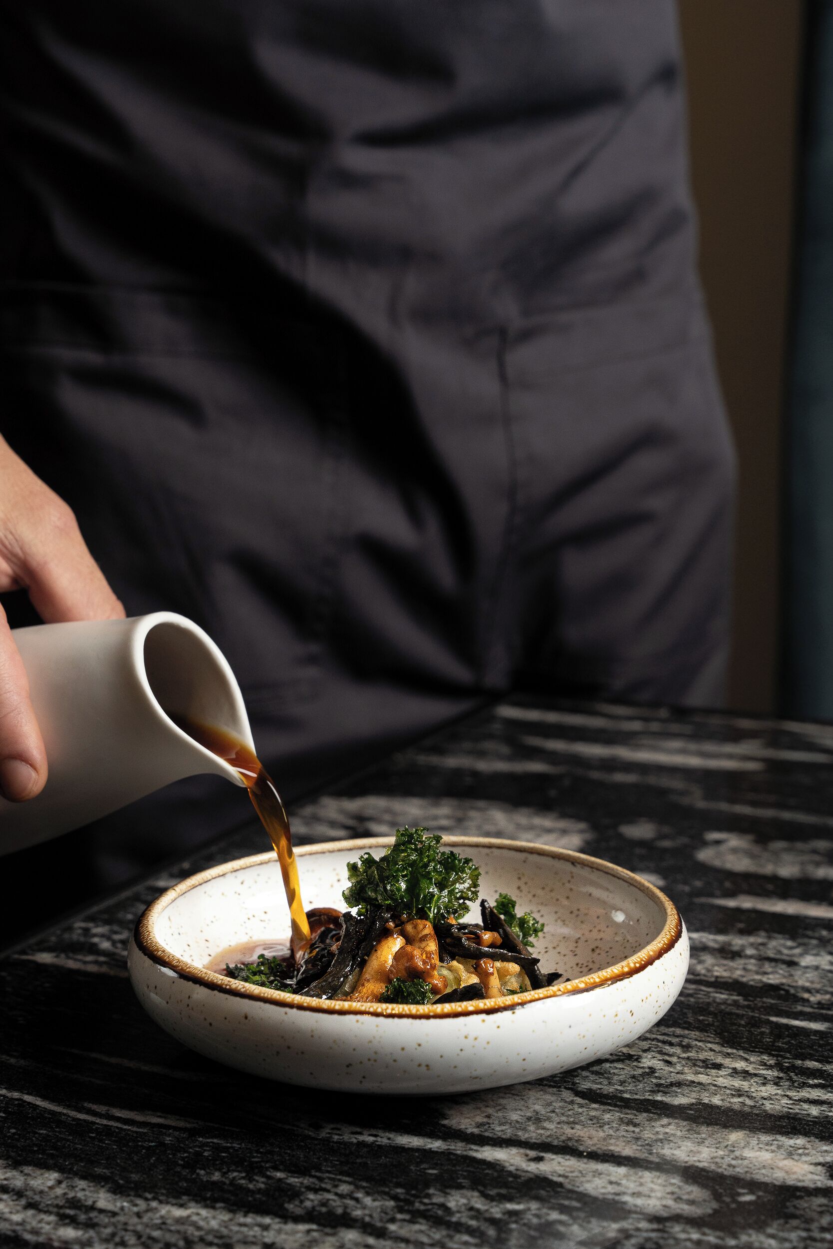 A person pouring a liquid from a white cup into a bowl of food on a marble table.
