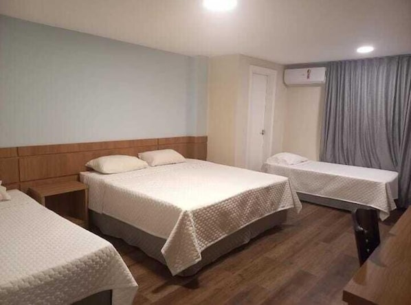 Quarto de hotel com duas camas de solteiro, uma cabeceira de madeira, uma pequena mesa de cabeceira, uma porta e uma cortina.