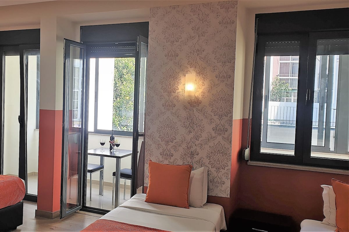 Habitación de hotel con dos camas, un balcón con mesa y sillas, y vistas al exterior.