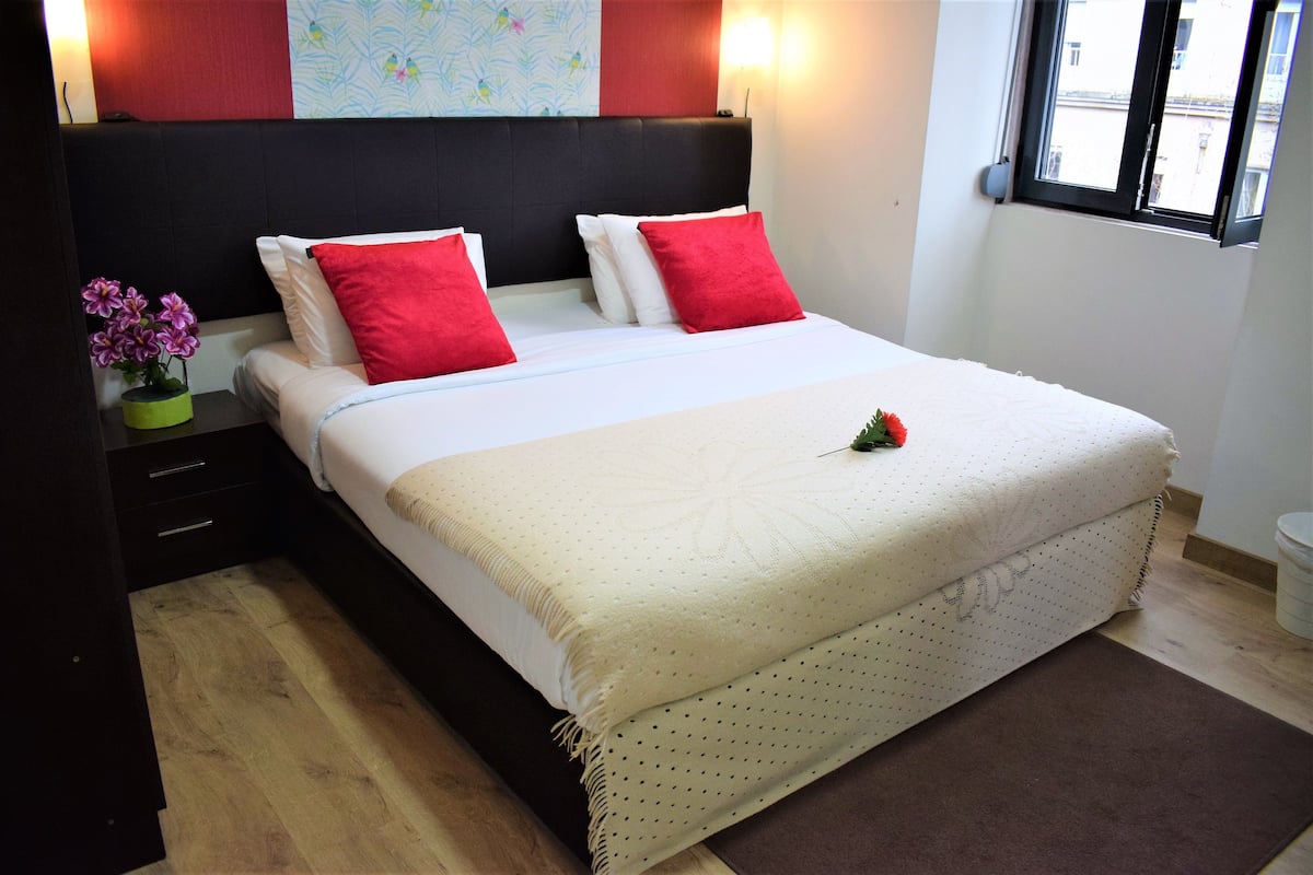 Una cama bien hecha con almohadas rojas y blancas, una alfombra marrón y una mesita con un jarrón de flores.