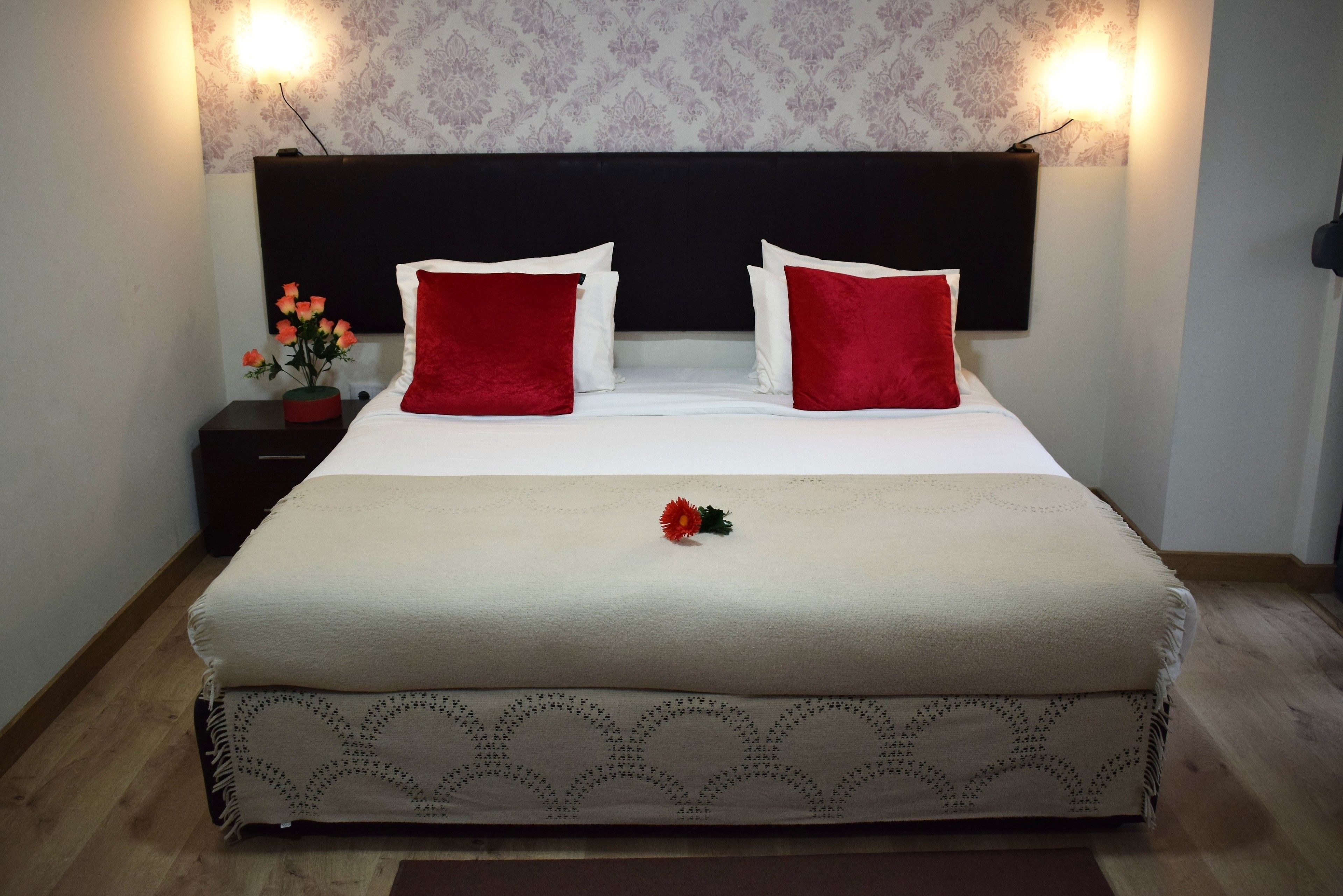 A neatly made bed with a white and beige bedspread, two red pillows, and a single red rose on the bed.