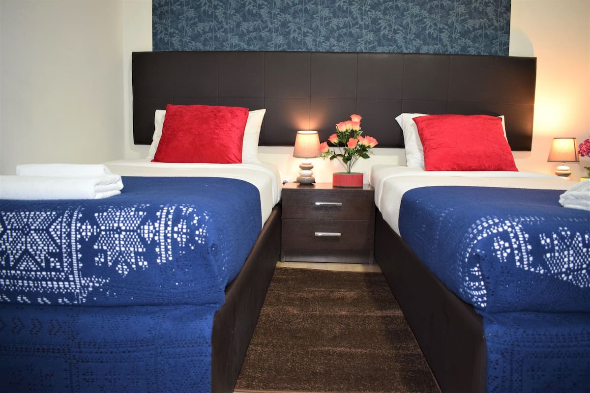 Habitación de hotel con dos camas, cada una con una colcha azul y almohadas rojas. Hay mesitas de noche con lámparas y un jarrón de flores.