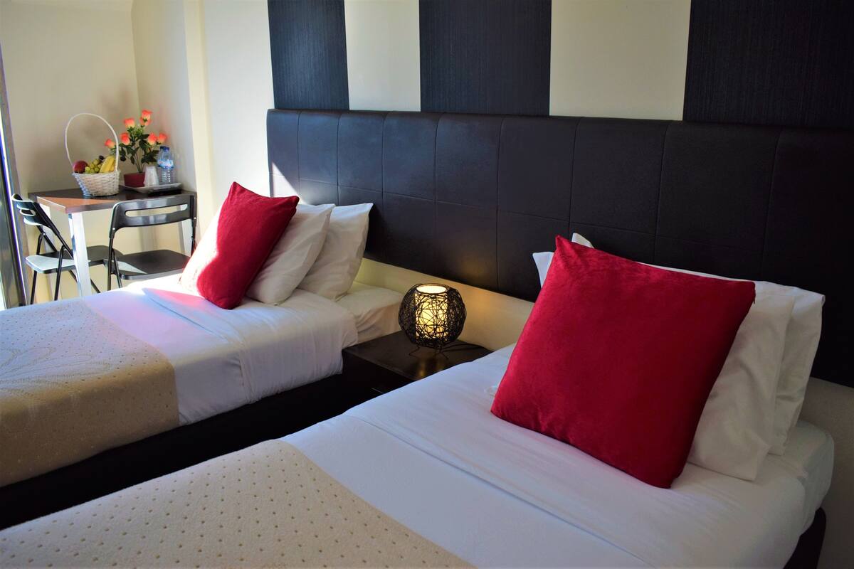 Habitación de hotel con dos camas, cada una con almohadas rojas y blancas, una mesita con una cesta de frutas y flores, y una mesita de noche con una lámpara.