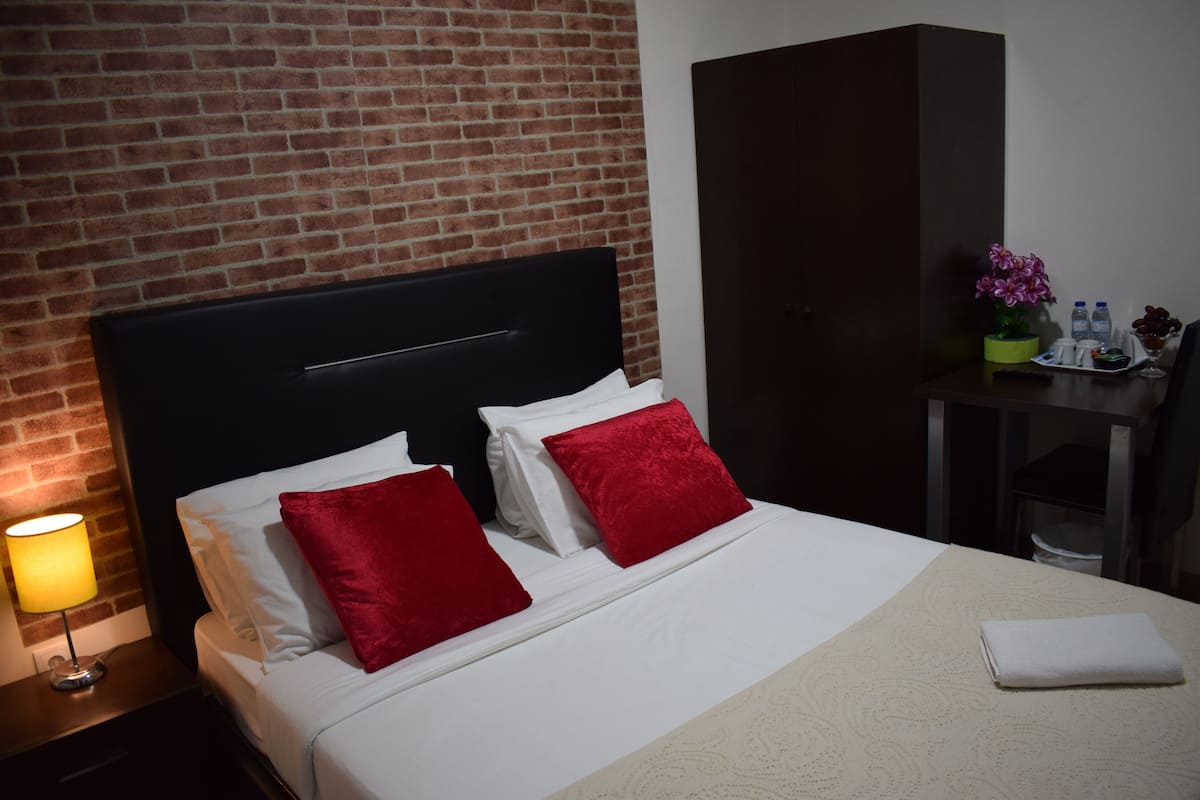 Habitación de hotel con pared de ladrillo, cabecero negro, ropa de cama blanca, almohadas rojas, una lámpara de noche, una mesita con un florero y una toalla doblada sobre la cama.
