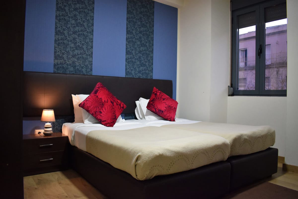Una cama bien hecha con una colcha color beige, dos almohadas rojas y un cabecero oscuro.