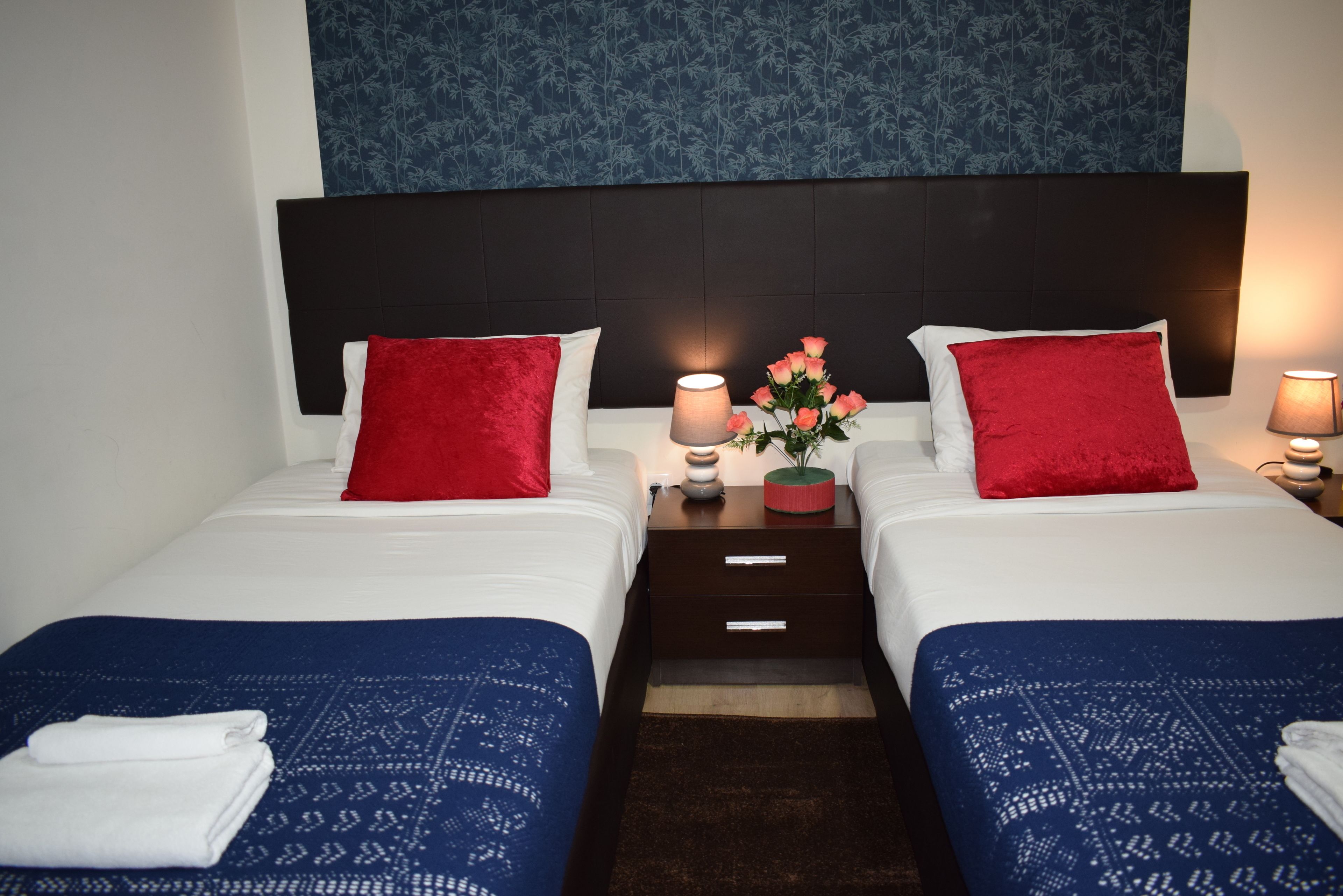 Een hotelkamer met twee bedden, elk voorzien van een rood kussen en een wit kussen, een nachtkastje met een lamp en een vaas met bloemen.
