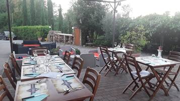 Un área de comedor al aire libre con mesas y sillas, rodeada de follaje y un muro de piedra.