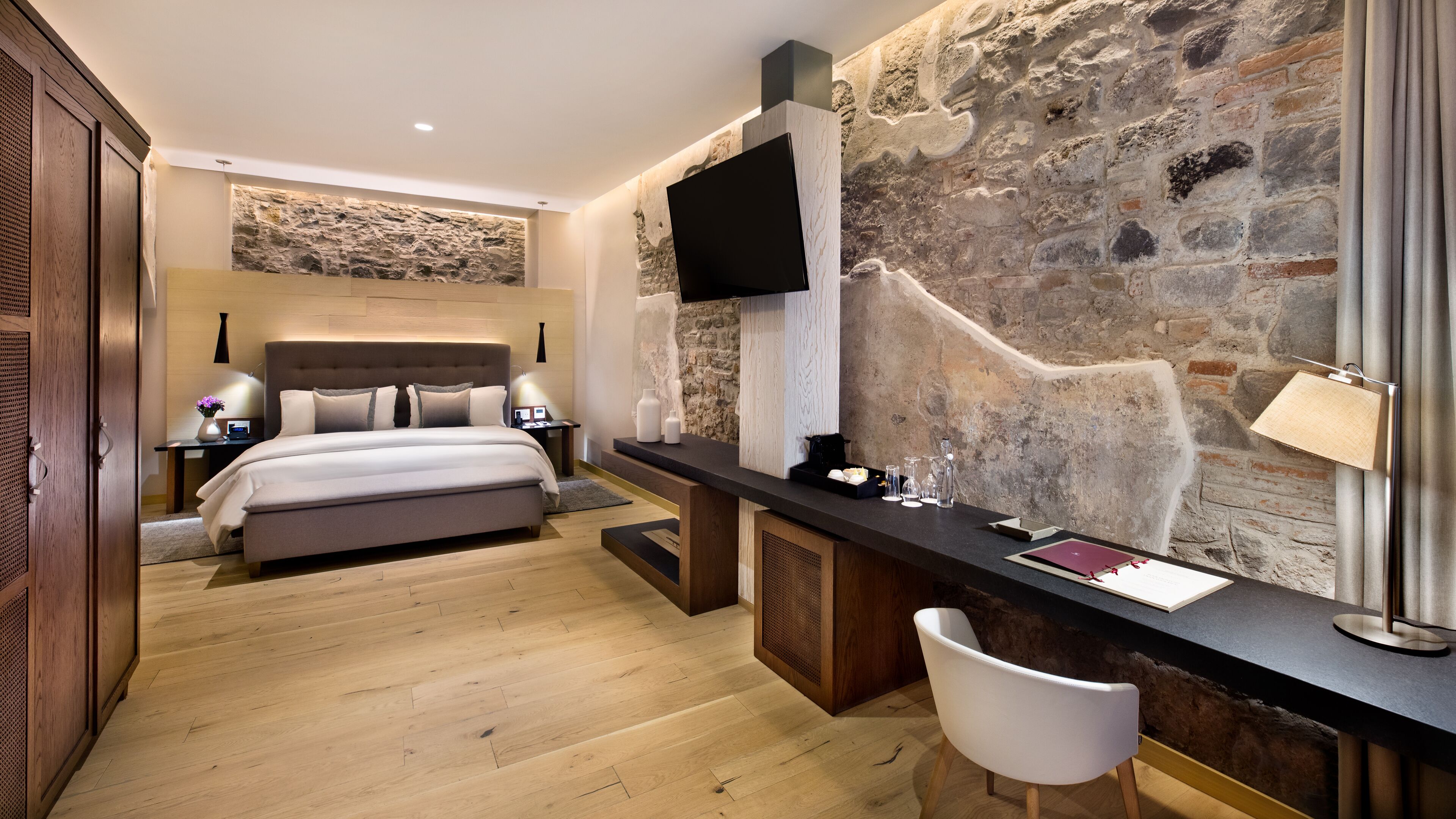 Un dormitorio moderno con una pared de acento de piedra, un televisor de pantalla plana, una mesita de noche con lámpara y una cama grande.