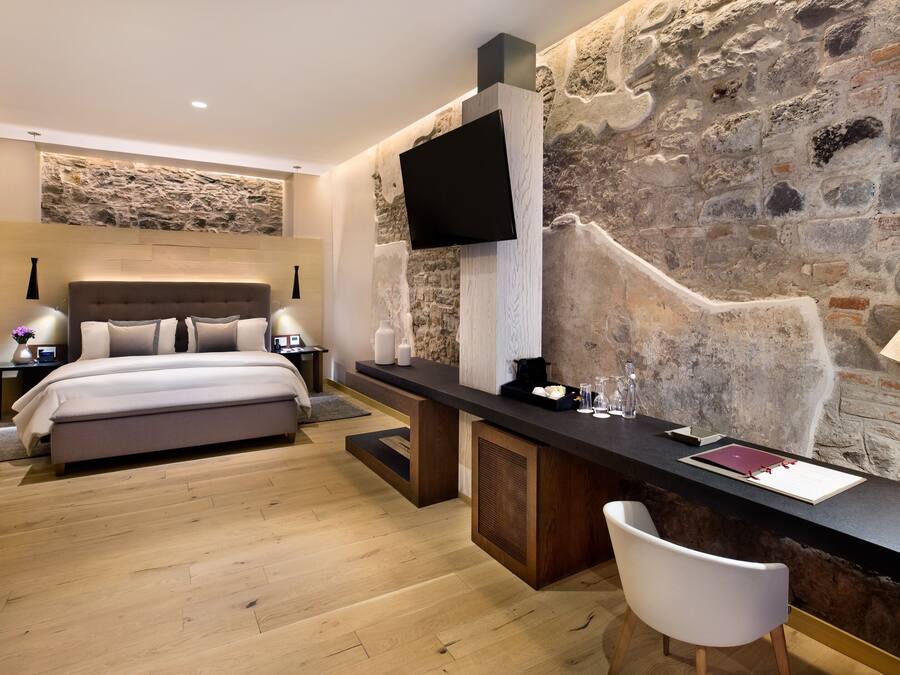 Un dormitorio moderno con una pared de acento de piedra, un televisor de pantalla plana, una mesita de noche con lámpara y una cama grande.