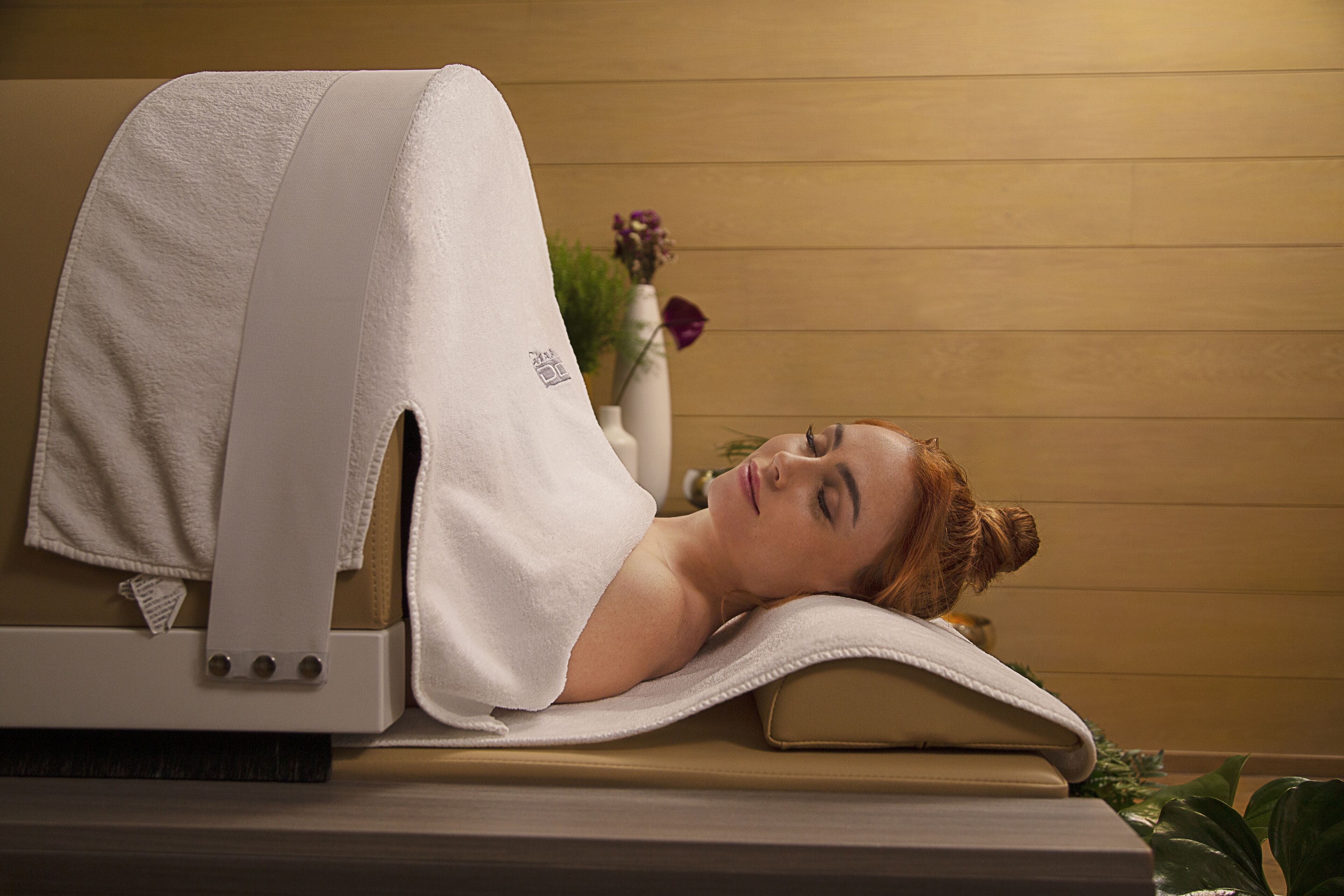 A person lying on a massage table with a towel draped over them, in a room with wooden paneling and a vase with flowers in the background.