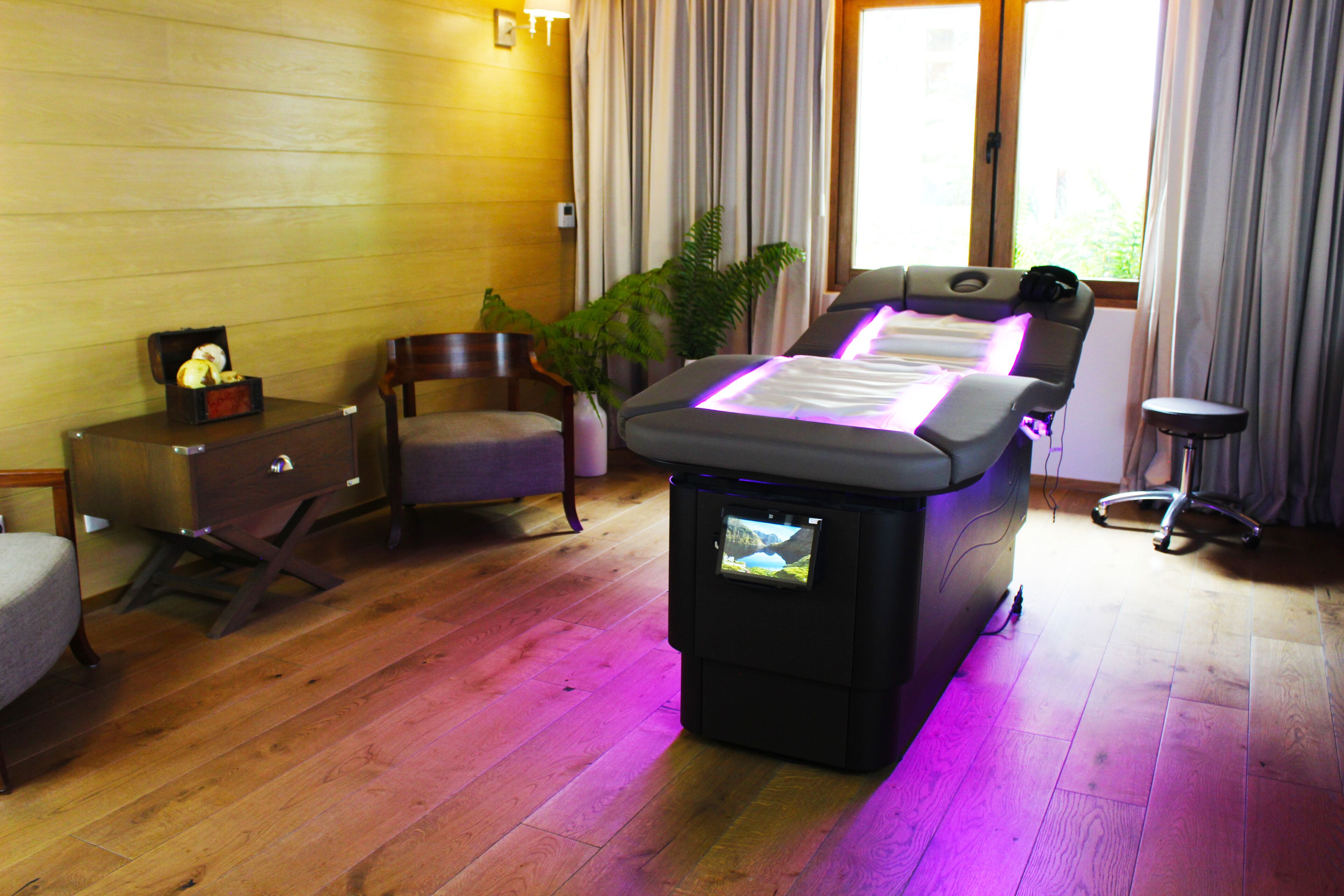 A massage therapy room with a massage table, a small cabinet, a chair, and a potted plant.