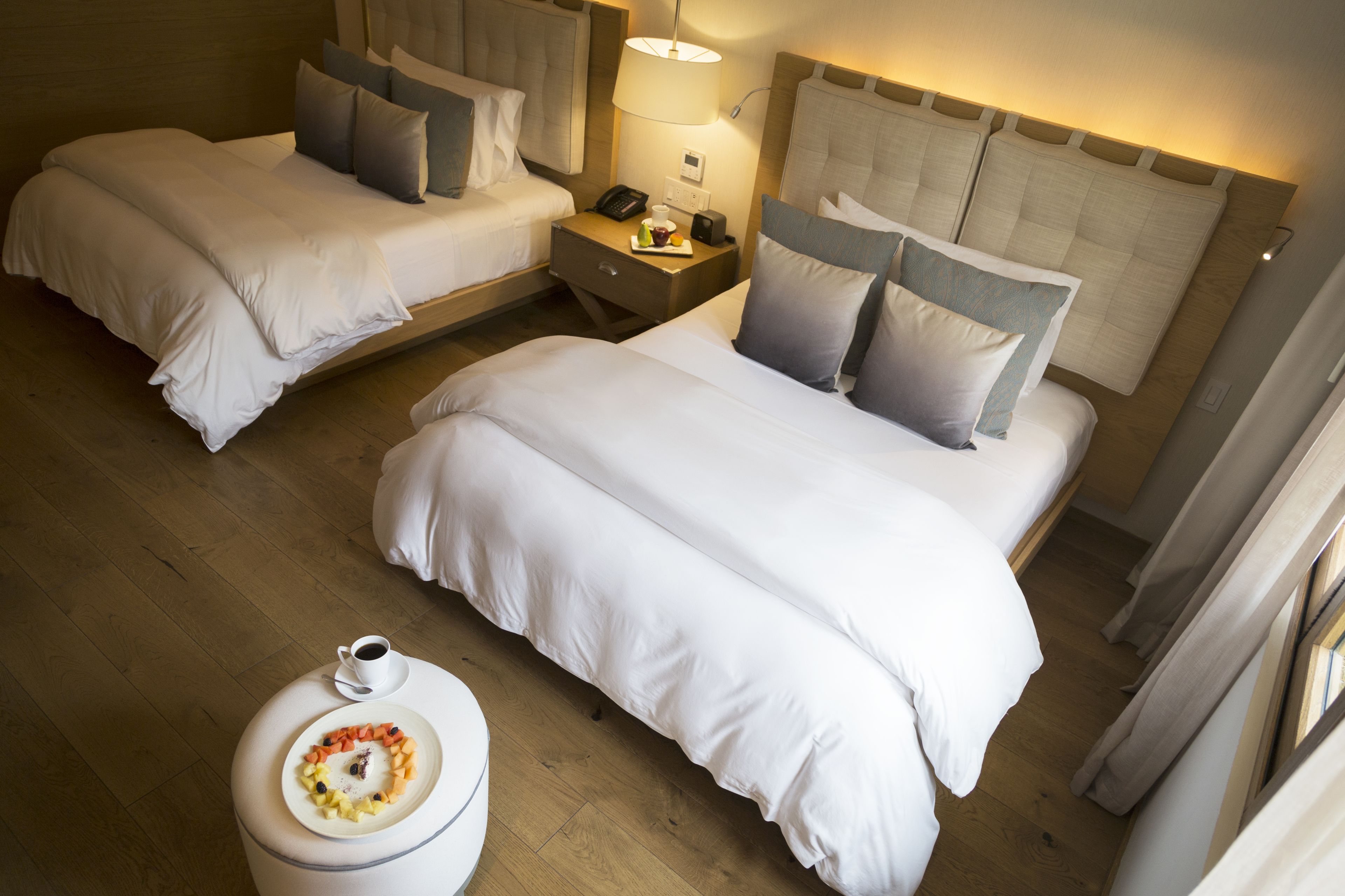A hotel room with two beds, a bedside table with a lamp, a small round table with a plate of food, and a window with curtains.