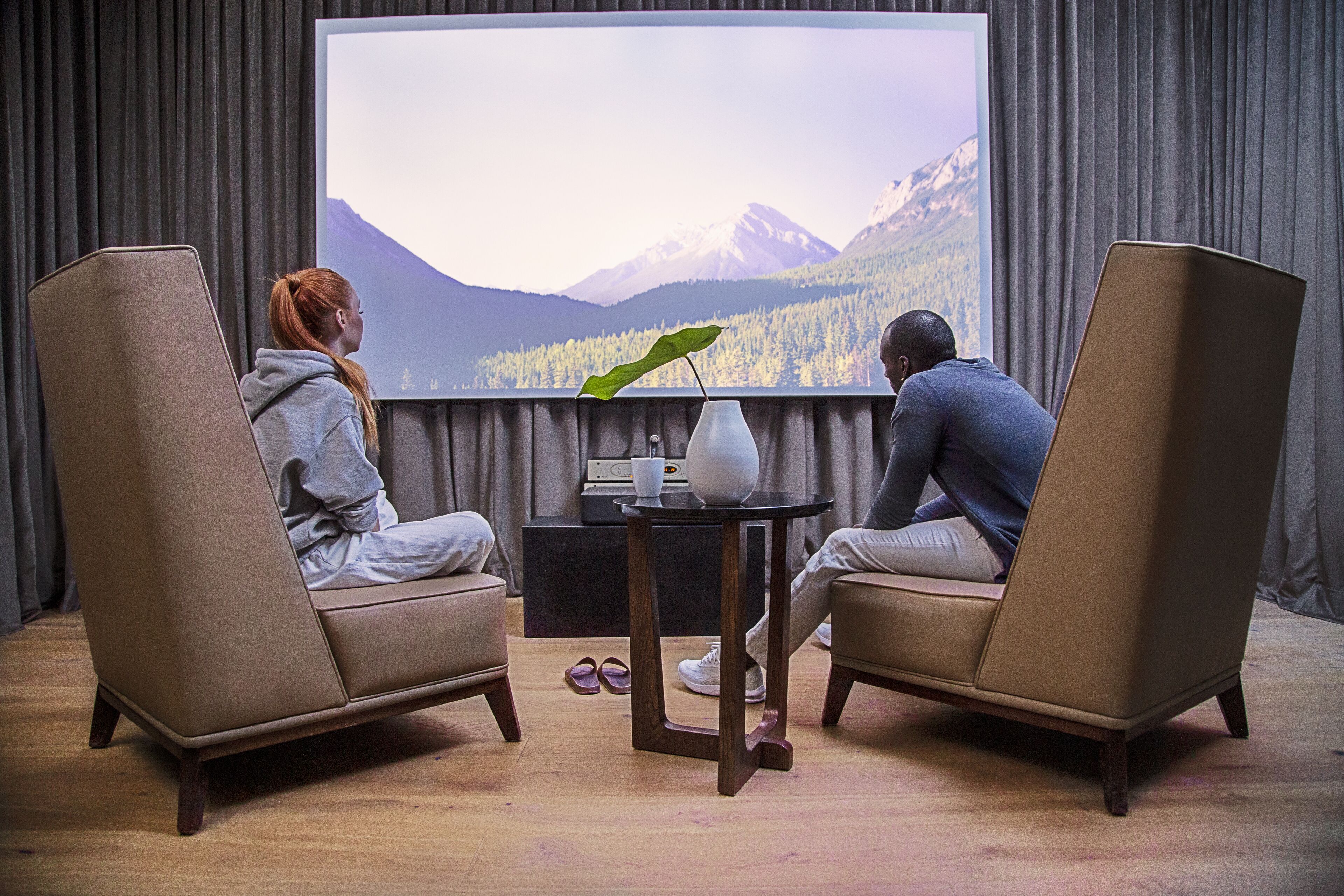 Dos personas sentadas en un sofá mirando una pantalla grande que muestra una vista panorámica de montañas.