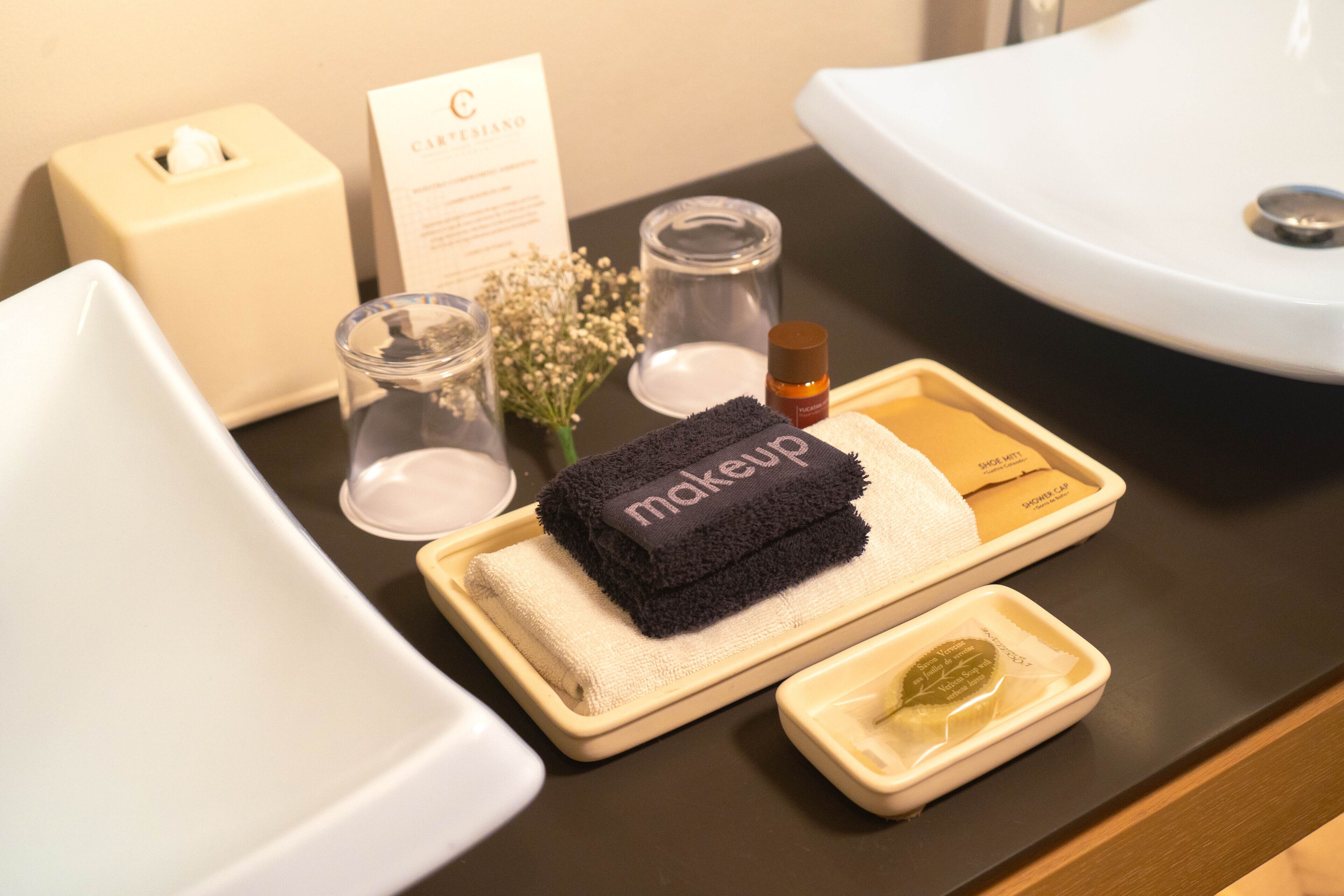 A bathroom counter with a makeup tray, a tissue box, a glass, and a small vase with flowers.