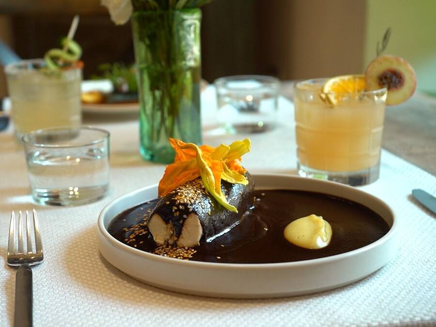 Un plato de postre con un manjar cubierto de chocolate, decorado con semillas de sésamo y pétalos de naranja, acompañado de una pequeña cucharada de crema.