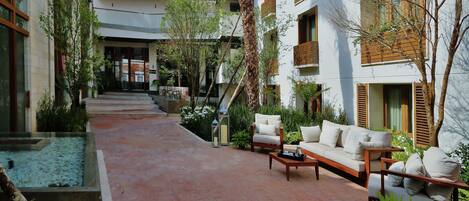 A courtyard with a pathway leading to a seating area with a sofa and coffee table. There are plants and a small water feature on the left.