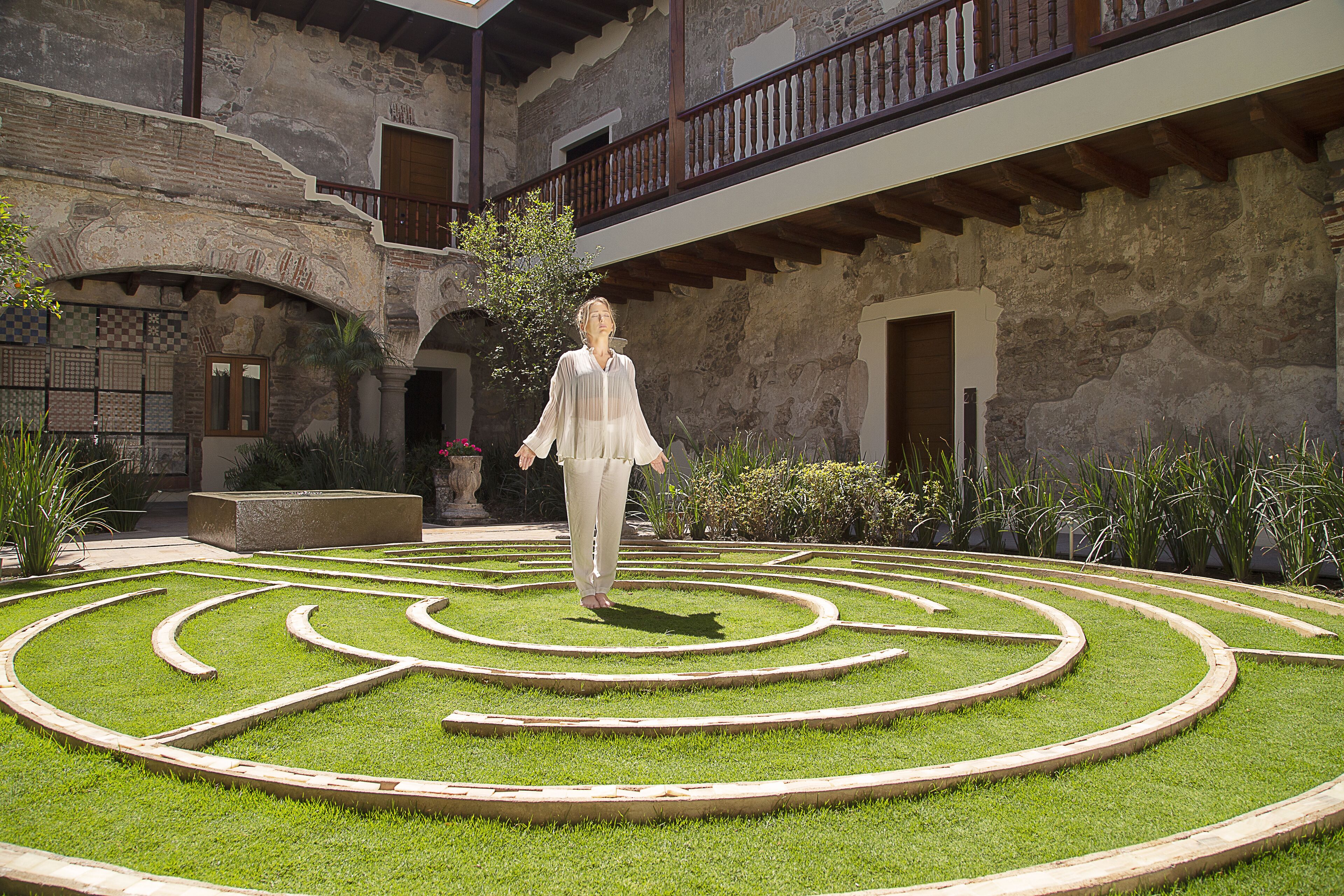 Un patio con un diseño geométrico de jardín, una persona caminando, y un edificio con muros de piedra y balcones de madera.