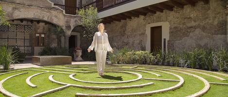 Un patio con un diseño geométrico de jardín, una persona caminando, y un edificio con muros de piedra y balcones de madera.