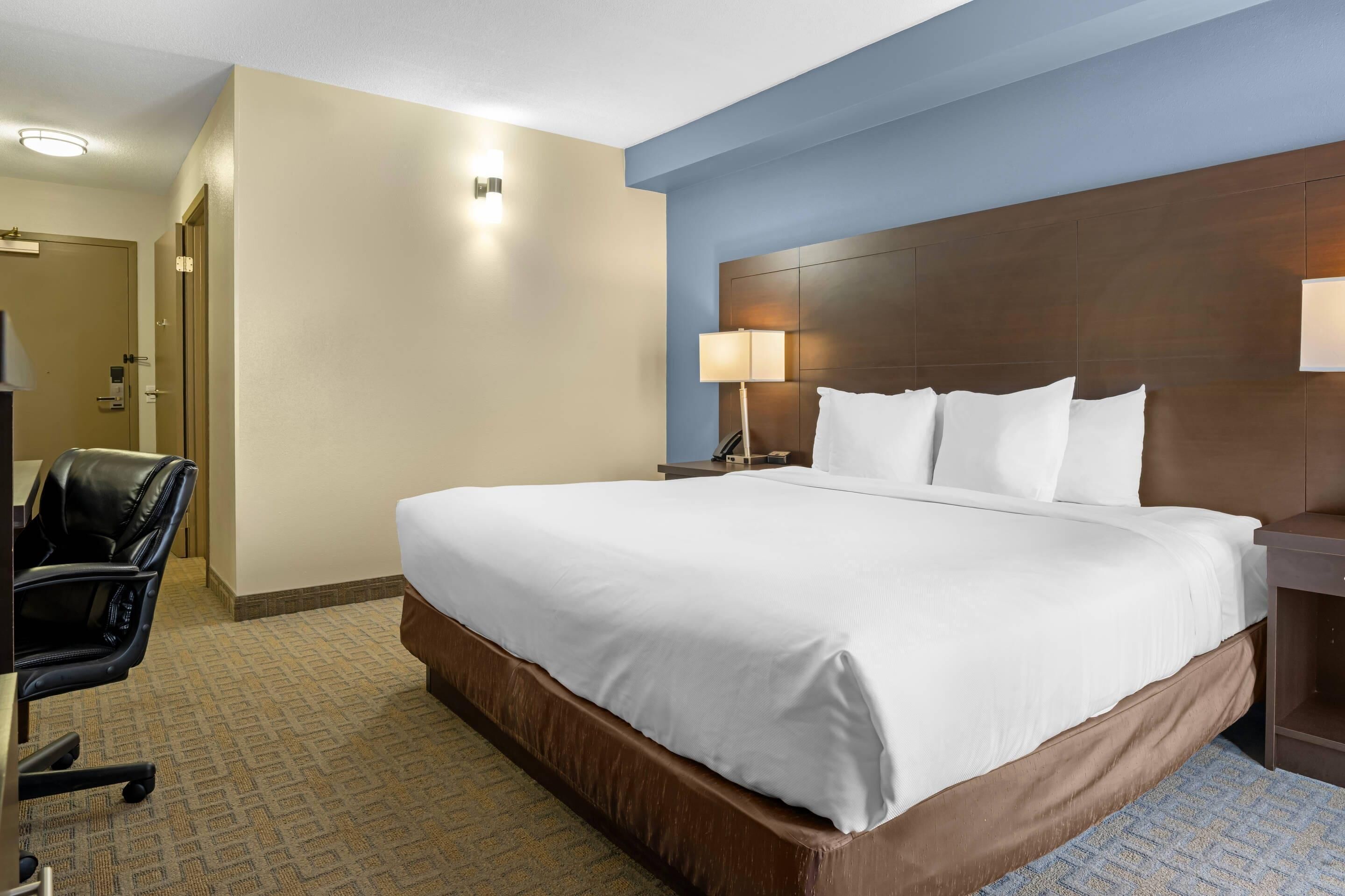 A hotel room with a large bed, a desk, a chair, and a nightstand with a lamp.