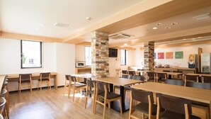 Un vaste espace dédié à la salle à manger, avec de nombreuses tables et chaises en bois, un pilier en pierre et, au fond, un coin cuisine.